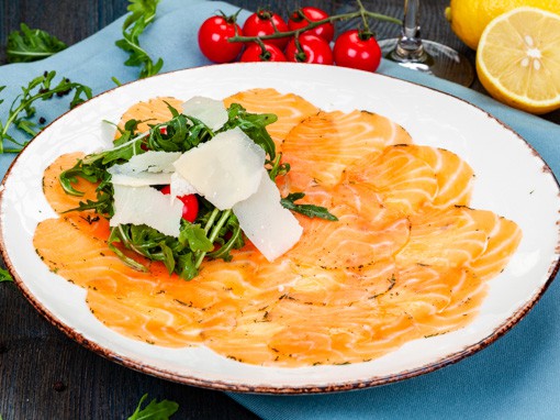 Das Gericht "Lachs-Carpaccio" hübsch dekoriert mit Rucola, Tomaten und Parmesan auf einem weißen Teller.