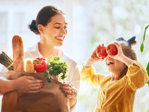 Stoffwechsel testen: Mutter hat Nahrungsmittel eingekauft, kleine Tochter hält sich zwei Tomaten lachend vor die Augen